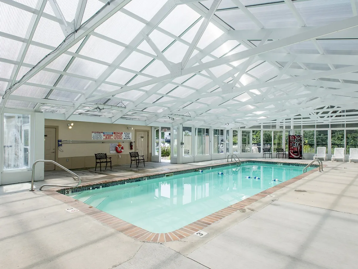 This image displays an inviting indoor pool area with white beams overhead and several lounge chairs arranged poolside. The tranquil blue water and spacious, bright setting provide a welcoming atmosphere for family swimming, relaxation, and socializing, regardless of the weather.