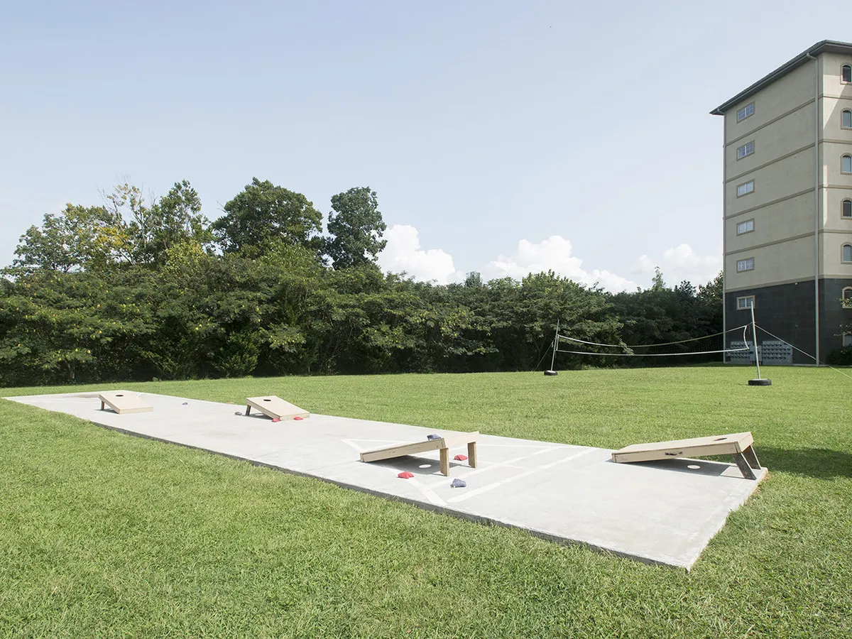 The image highlights a vibrant grassy area equipped for both play and relaxation, featuring cornhole boards, a volleyball net, picnic tables, and space for socializing. The backdrop of trees and a building ensures a safe, contained environment for active recreation.