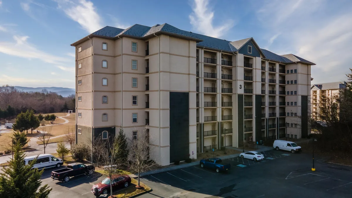 This image captures the exterior of a four-story building surrounded by trees, with a parking lot in the foreground. The setting is calm and inviting, with hints of the nearby mountains visible in the background. The space around the building suggests areas for gathering and easy access for groups arriving together.