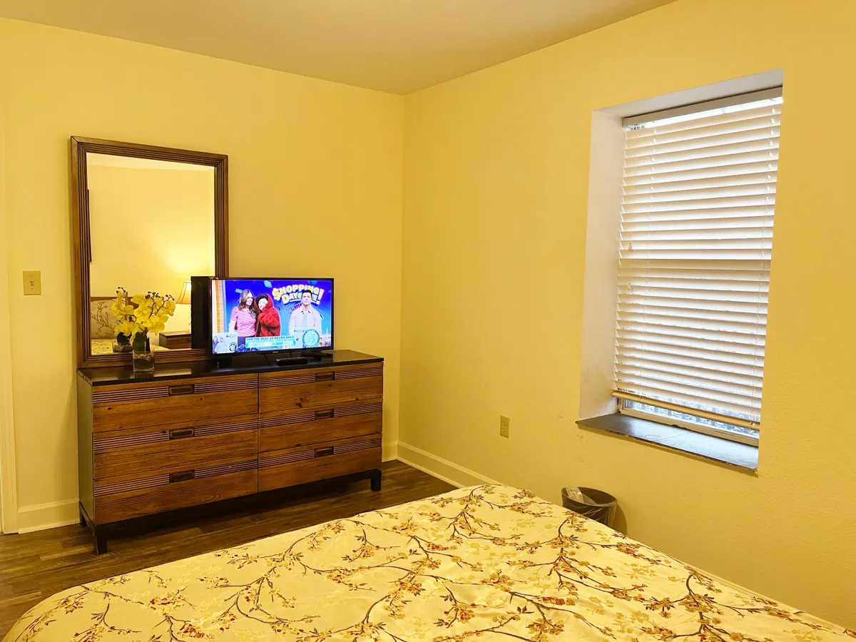 A cozy bedroom with floral bedding, a television atop a dresser with a mirror, and a window with adjustable blinds. The space is thoughtfully appointed for comfort, offering both relaxation in bed and the convenience of enjoying favorite shows privately.