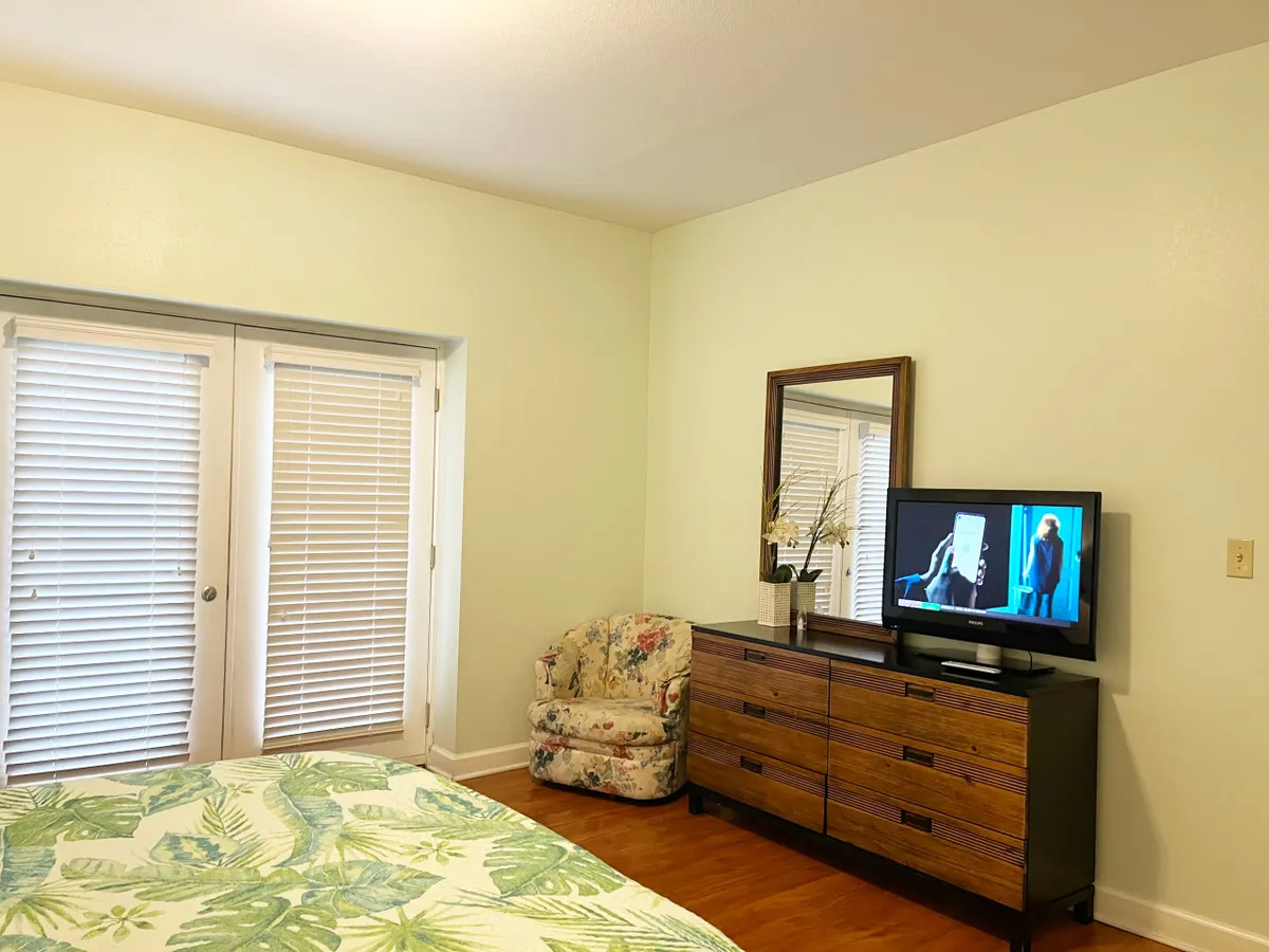 A tranquil bedroom scene with a floral-bedded bed, dressing mirror, television, and an armchair by the window. The room is inviting, perfect for both restful sleep and peaceful personal time.