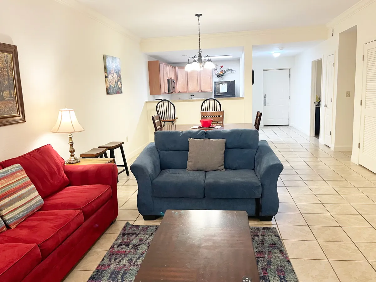 A cozy, open living and dining area with a red sofa, blue sofa, dining table with chairs, decorative lamp, and art on the wall. The space is arranged to encourage group interactions—whether for movie nights, meals, or casual conversations—with plenty of soft seating and a welcoming atmosphere.