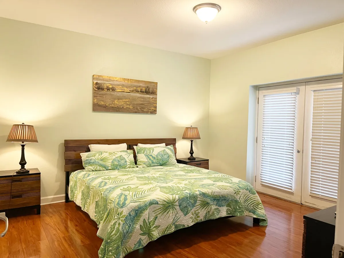 A tranquil bedroom featuring a king or queen bed adorned with floral bedding, flanked by matching bedside tables with elegant lamps. Soft morning light pours in through French doors, illuminating tasteful wall art and creating an inviting atmosphere for rest and relaxation.