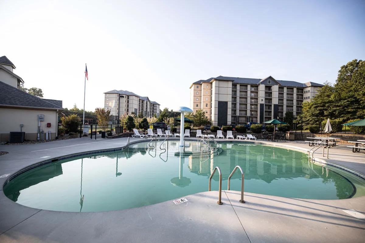The image features a sparkling swimming pool bordered by lounge chairs, with green trees and resort buildings in the background. The relaxed mood invites families to unwind poolside or enjoy social events in a resort setting.