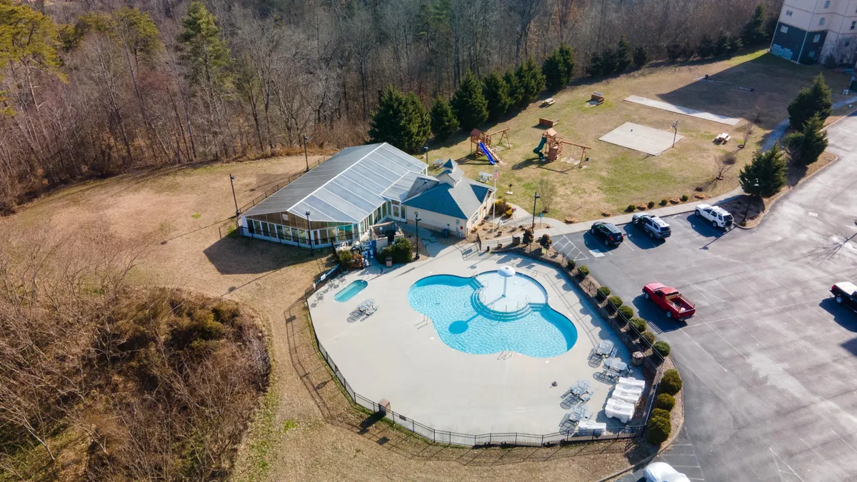 This vibrant exterior shot showcases a large swimming pool surrounded by a recreational area, visible playground equipment, and a nearby community center. The scene is lively and inviting, suggesting a family-friendly atmosphere where kids can play and parents can relax close by.